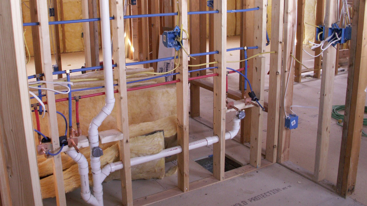 Exposed wall framing in a house under construction, showing plumbing pipes, electrical wiring, and insulation between wooden studs.