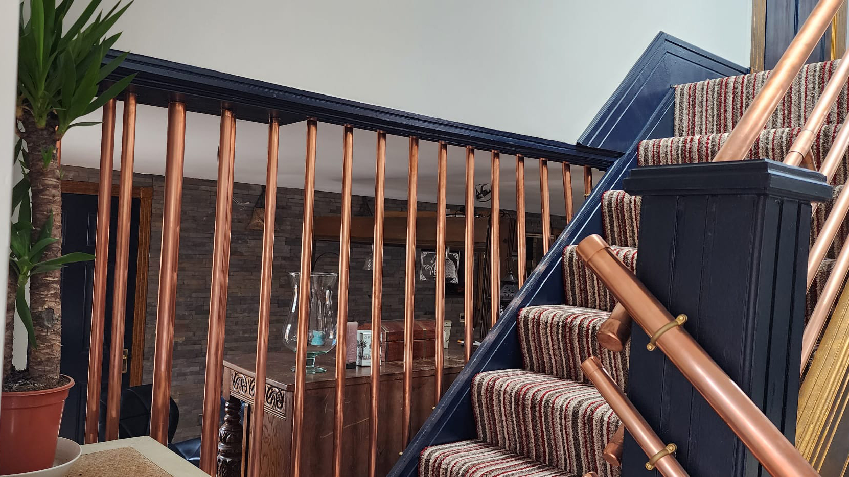 Blue skirting board with a staircase covered in lined coloured carpet with copper handrails, with view of the view of the neighbouring room.